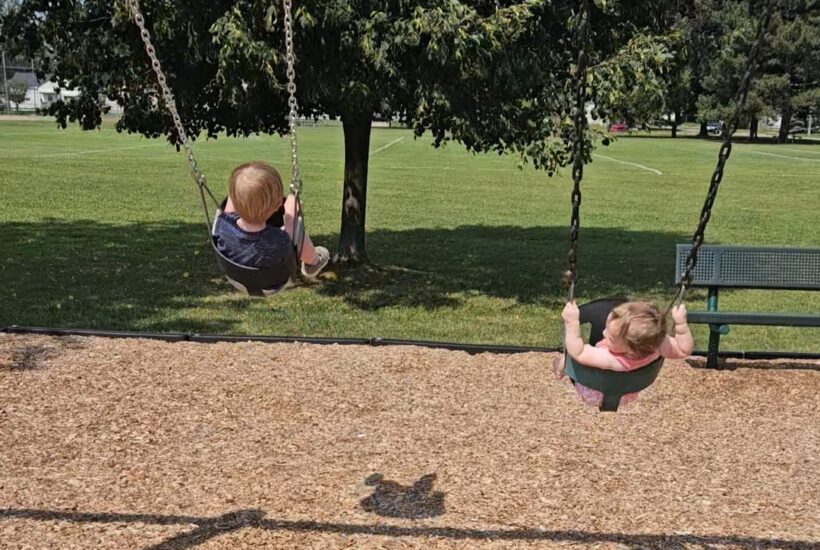 two children on swings at the park with a grassy field in the background